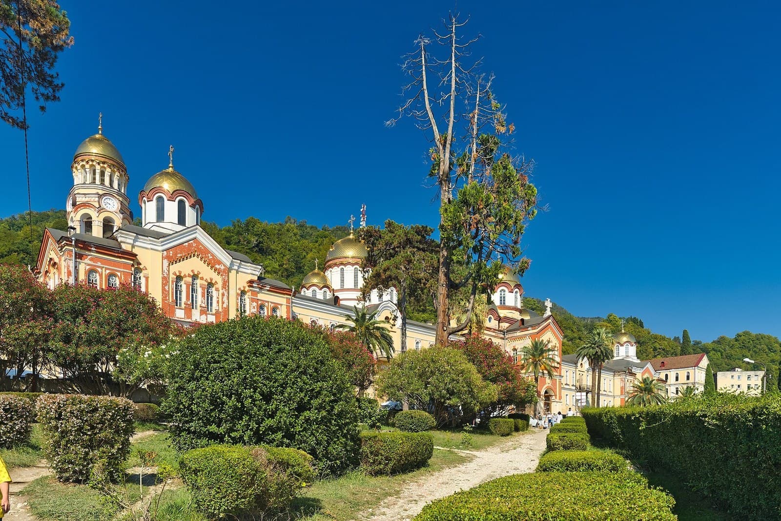 The gilded domes and ochre and red brick facades of the New Athos monastery on a wooded hillside above Novy Afon, with clipped hedges and a gravel path in the foreground