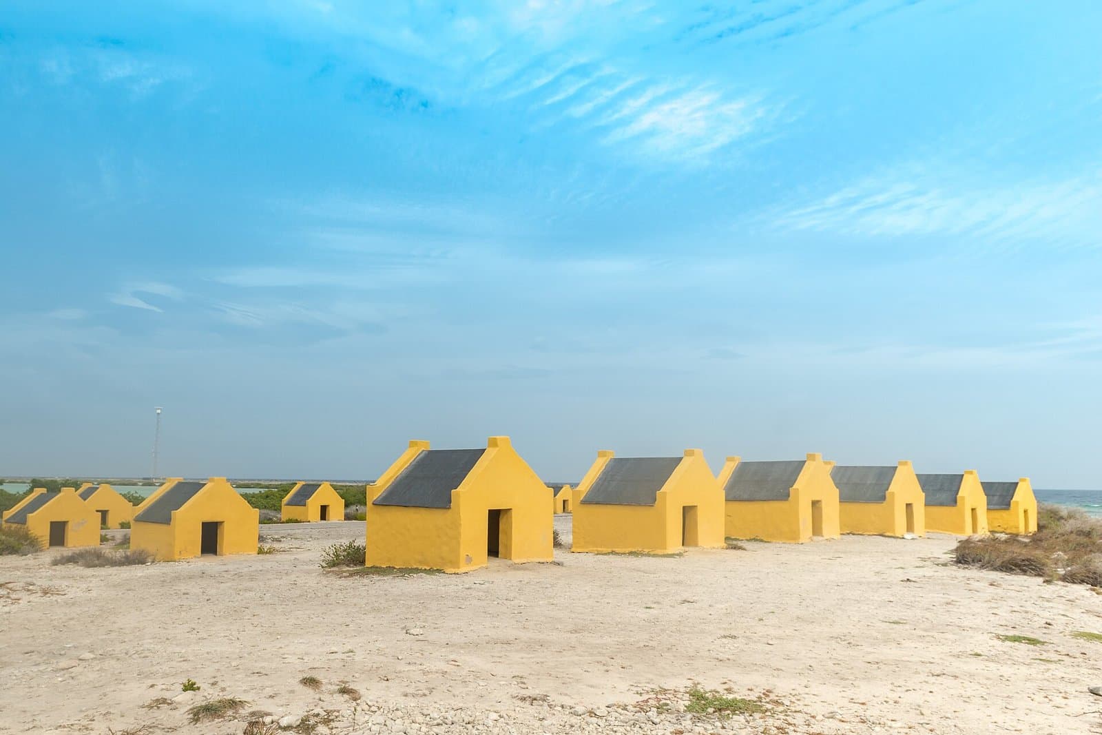 A row of small gabled stone huts painted ochre yellow with dark slate roofs, standing on white coral gravel beside the salt pans on the south coast of Bonaire, with low scrub and open sea behind them