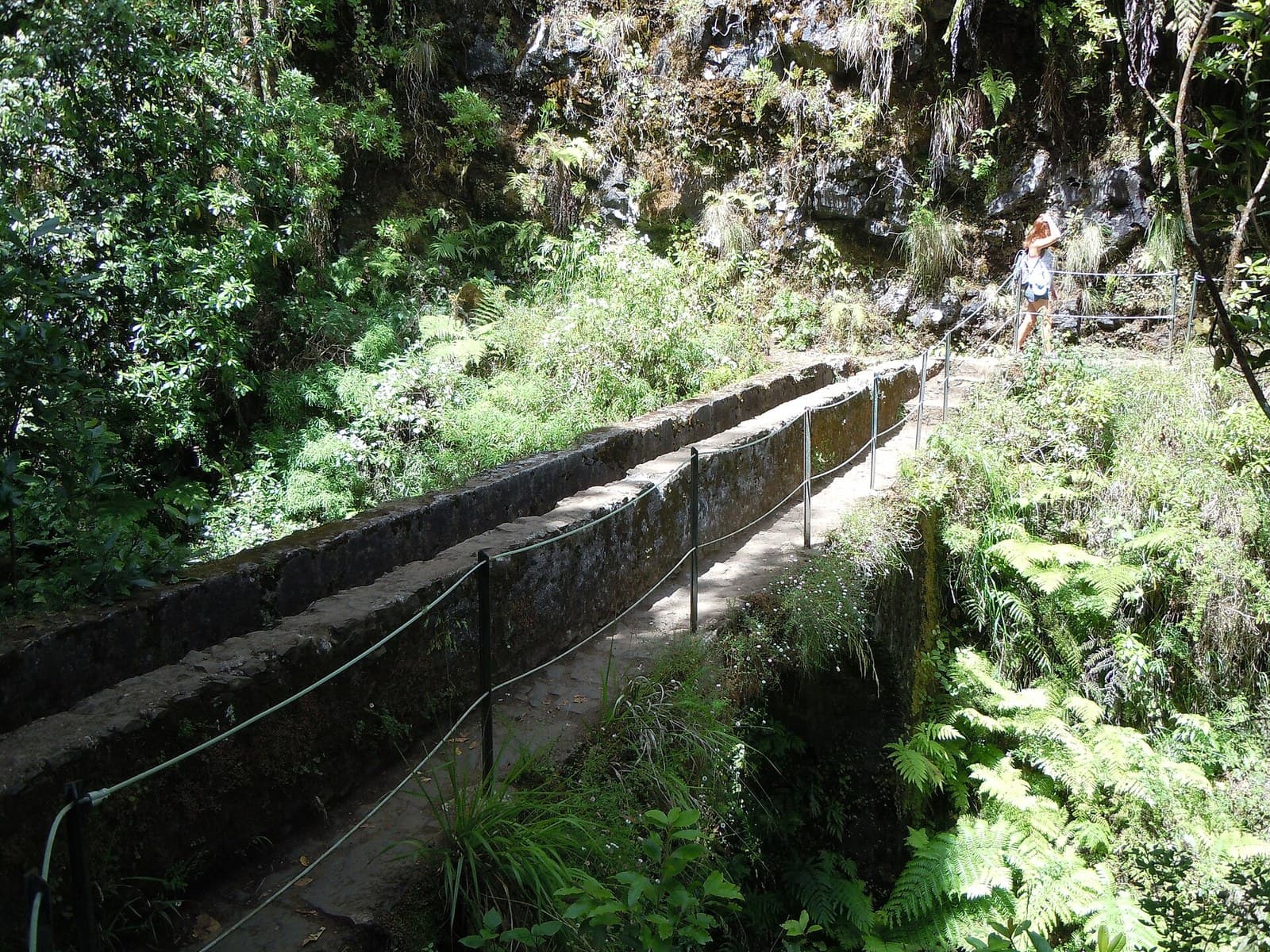 A stone-lined levada channel running along a hillside on the Levada do Caldeirão Verde above Santana, Madeira, with a narrow walking path beside it, a rope handrail and laurel forest and ferns on both sides