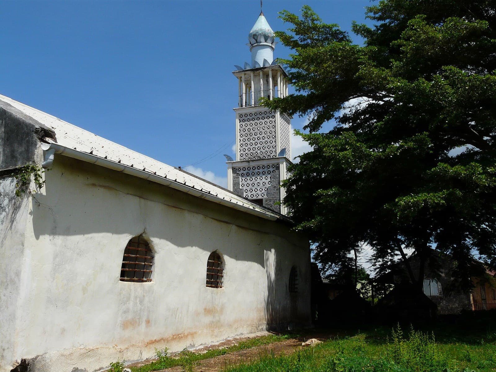 The old Friday mosque at Tsingoni, Mayotte: a long whitewashed prayer hall with barred windows under a sloping tin roof, and behind it a tall minaret of pierced white screens topped by a small dome