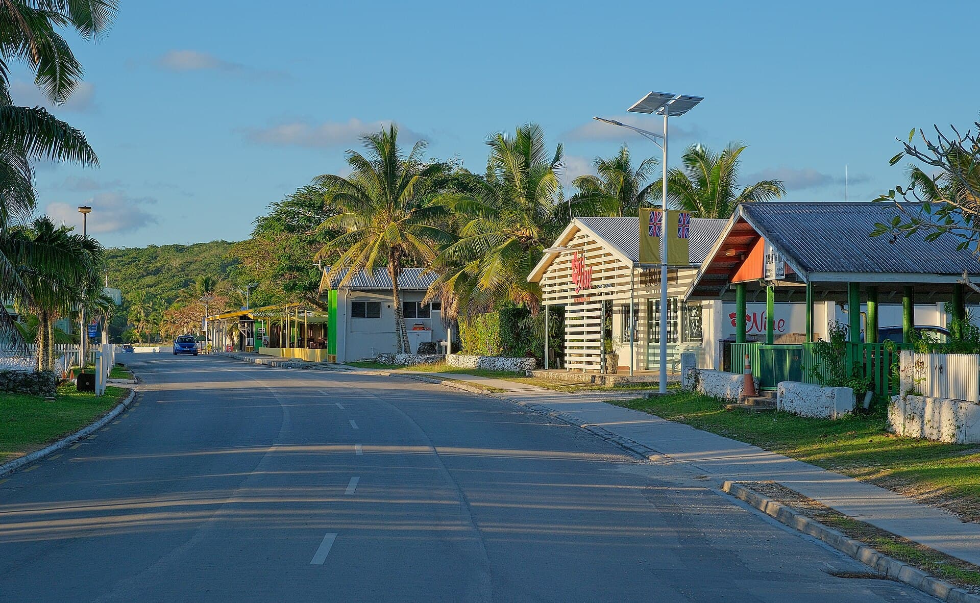 The main street of Alofi, Niue, in late afternoon, with single-storey shops, a solar-powered street light and coconut palms along an almost empty road