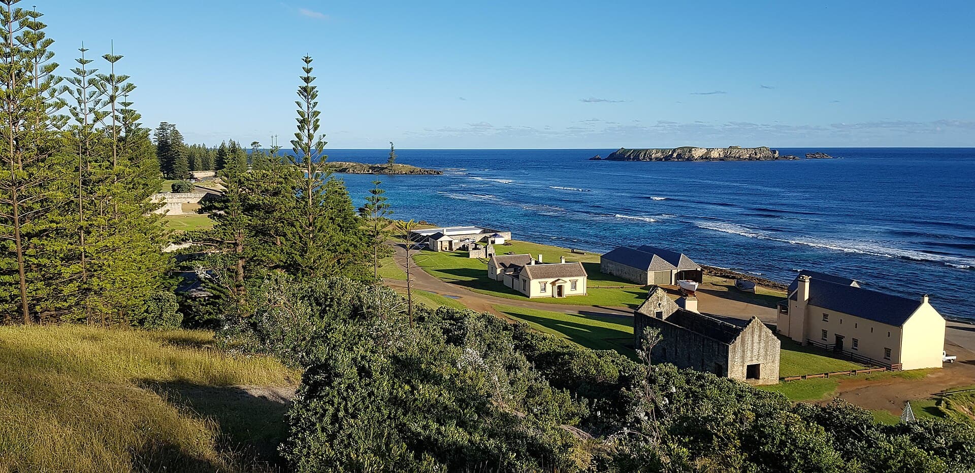 The Georgian stone and timber buildings of Kingston on the grass above a reef-fringed bay, with Norfolk pines in the foreground and a low offshore island on the horizon