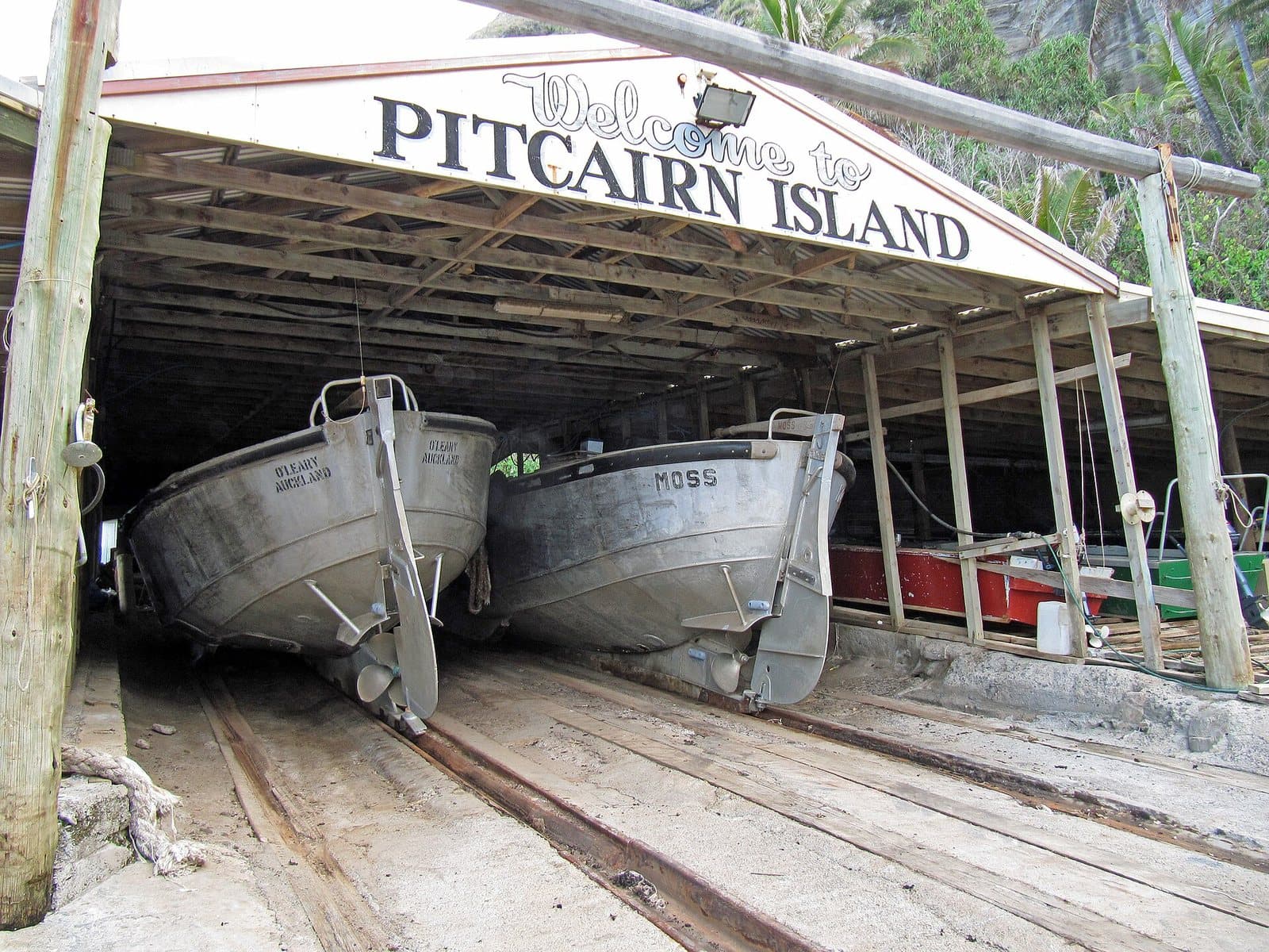 Two aluminium longboats on rails inside the open-fronted boat shed at Bounty Bay, Pitcairn Island, under a painted sign reading Welcome to Pitcairn Island
