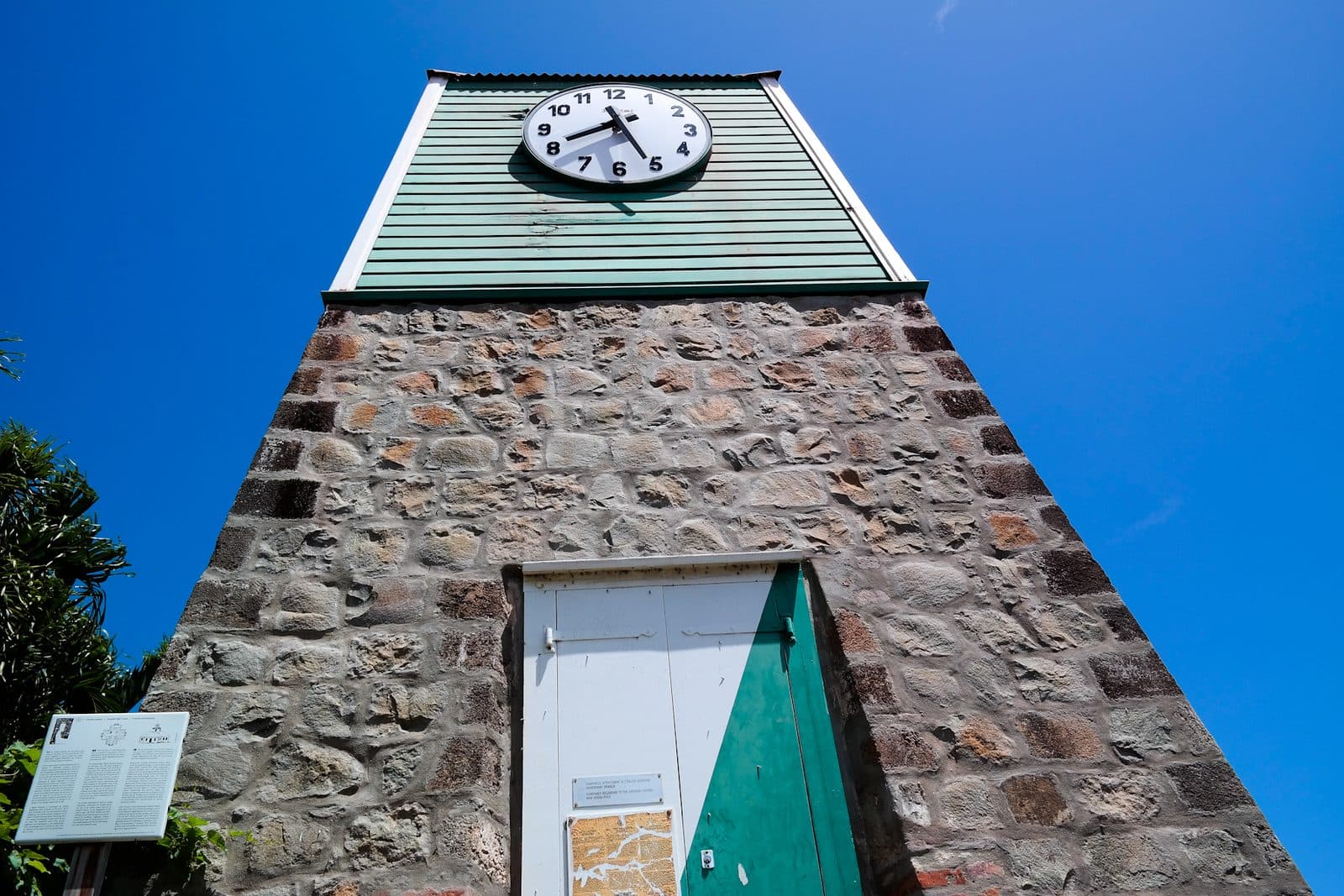 The Swedish bell tower in Gustavia seen from below against a clear sky, a square tower of rough grey and ochre stone with a white-framed door and a green weatherboarded upper stage carrying a white clock face