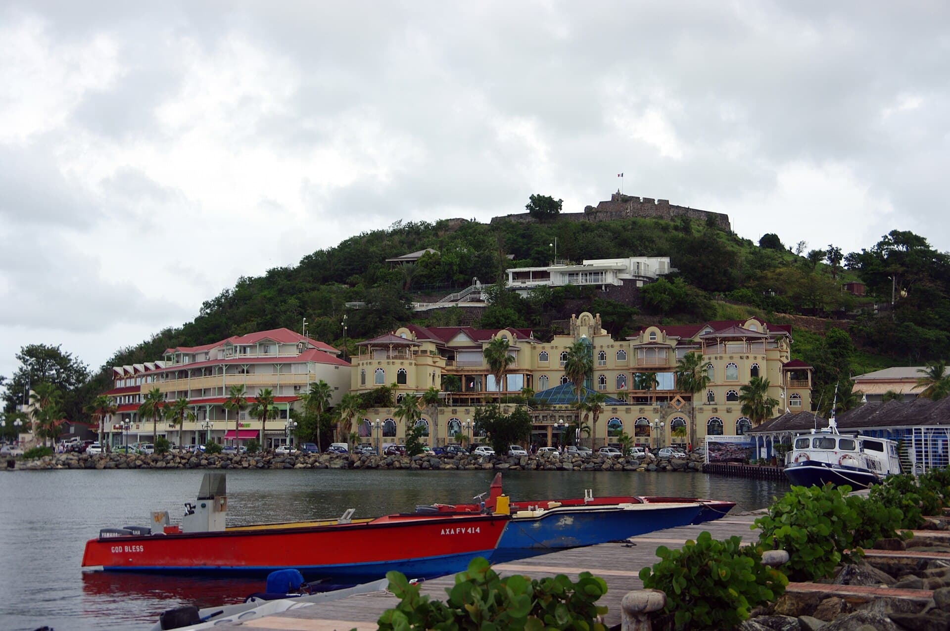 The harbour at Marigot on the French side of Saint Martin, with open fishing boats at the quay, creole-style waterfront buildings with balconies and red roofs, and the stone ruins of Fort Louis flying the French flag on the hill above