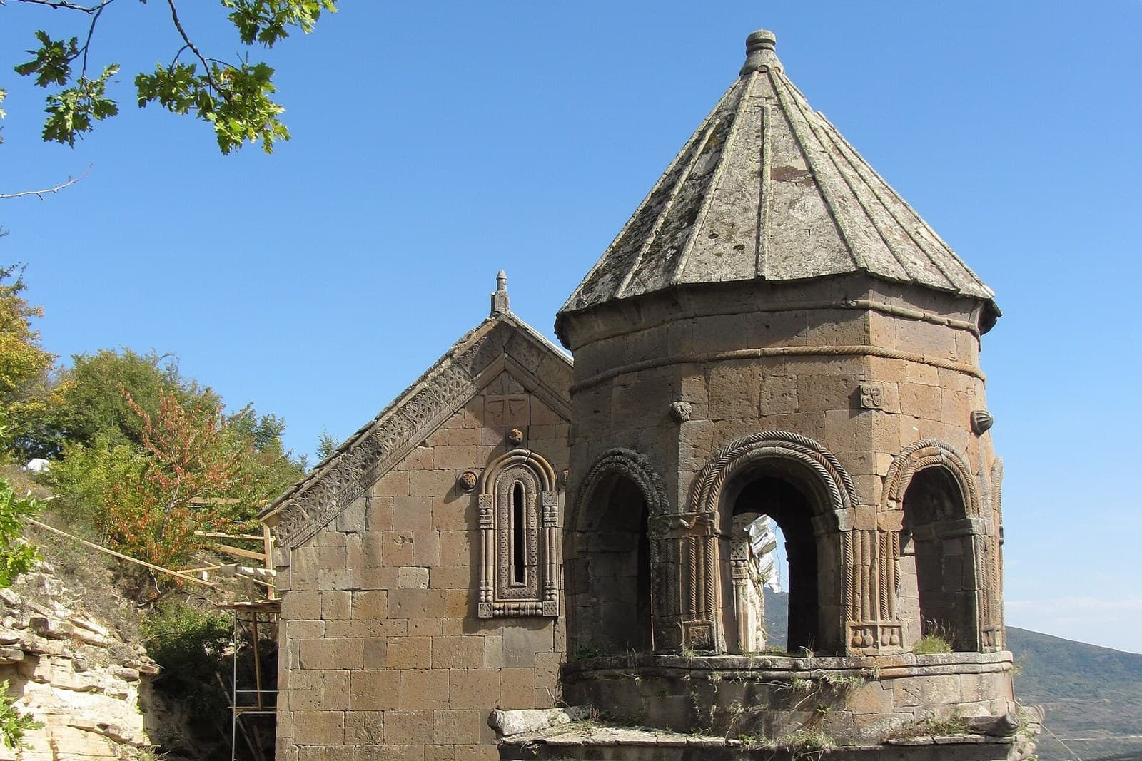 The carved stone drum and conical roof of the medieval church at Tiri monastery near Tskhinvali, with blockwork walls, an ornamented window and wooded hills behind