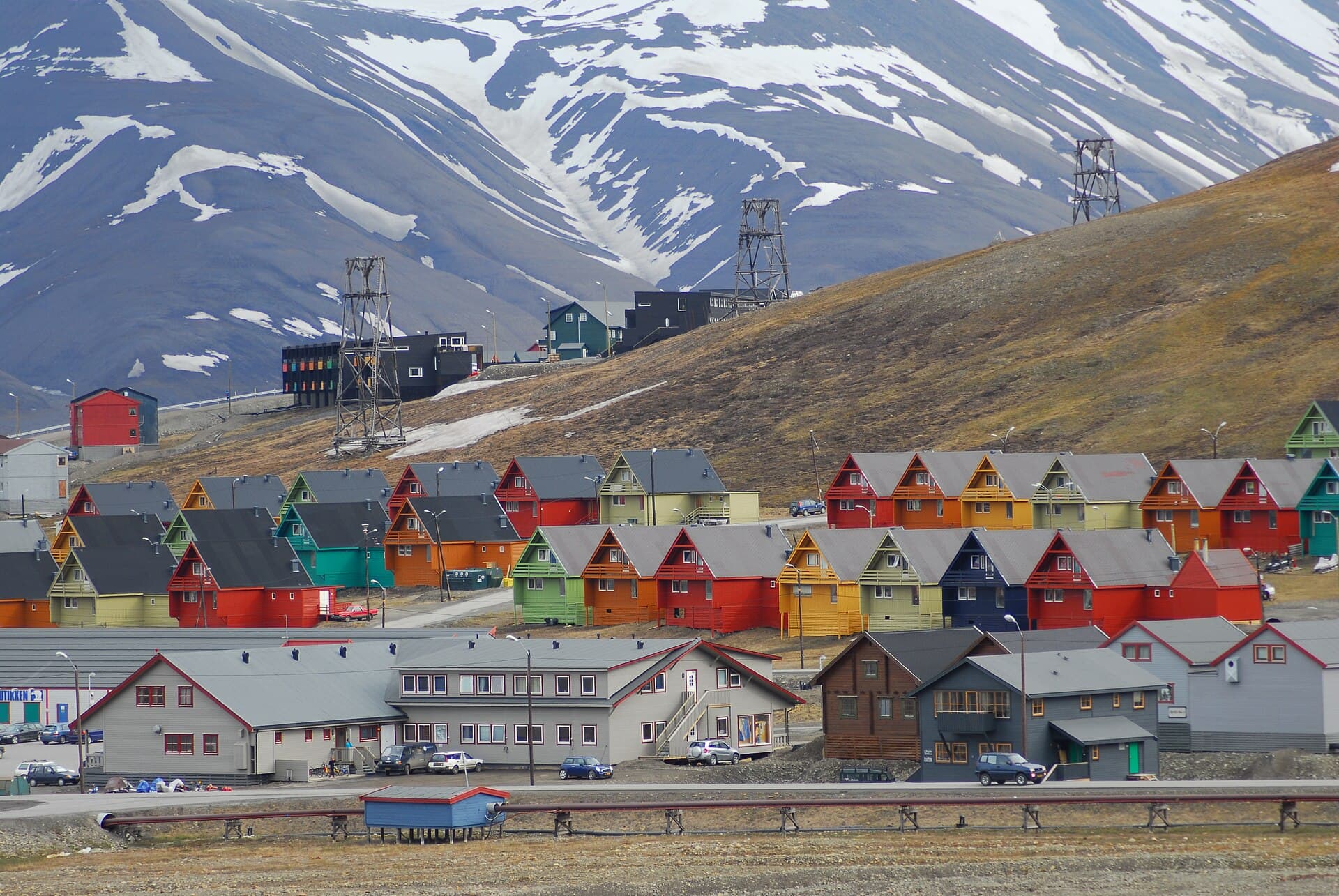 Rows of brightly painted timber houses in Longyearbyen below a snow-streaked mountain, with the steel trestles of the old coal cableway standing on the slope behind