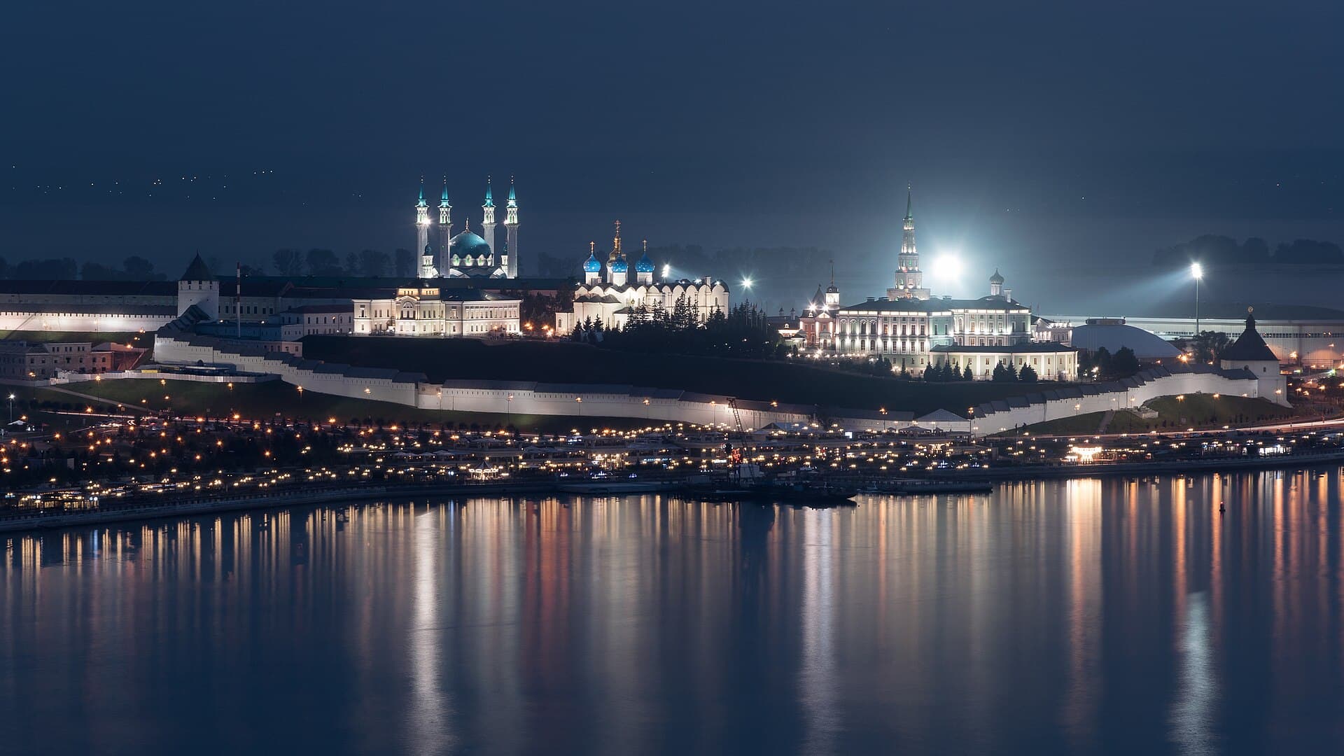 The Kazan Kremlin at night across the Kazanka river, with the lit minarets and blue dome of the Qol Sharif mosque and the domes of the Annunciation Cathedral inside the same white walls