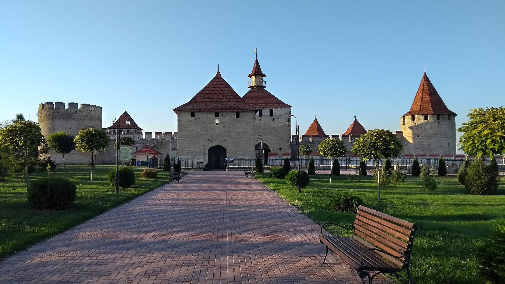 The south side of the Bender fortress, its pale stone curtain wall and cone-roofed towers rising above a park with clipped shrubs and a brick path