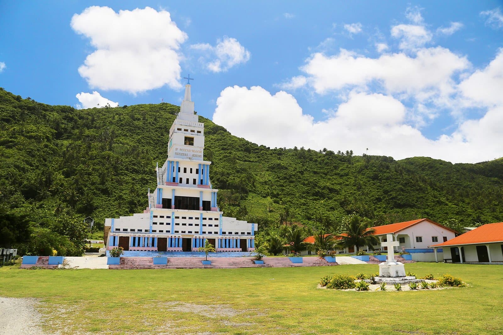 The stepped white and blue tower of the Basilica of Saint Peter Chanel at Poi on Futuna, standing on mown grass below a forested ridge, with a white stone cross to the right