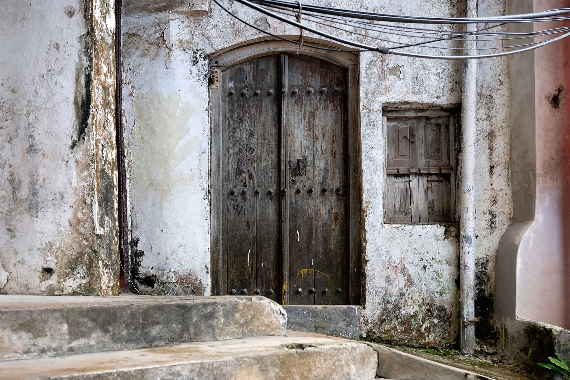 A studded wooden door with an arched frame set into a weathered coral-stone wall in Stone Town, with a shuttered window beside it and stone steps in the foreground