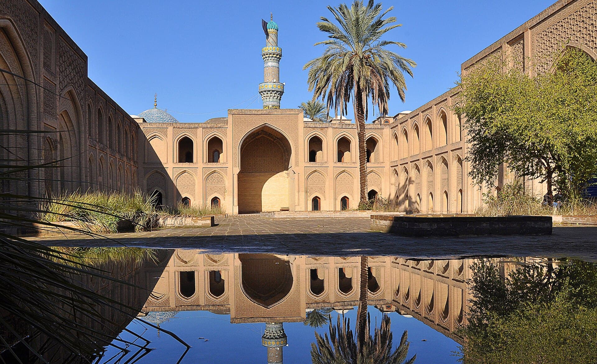 The brick courtyard of the thirteenth-century Mustansiriya Madrasa in Baghdad, its arcades and the minaret behind reflected in a long pool