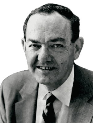 A black-and-white portrait of a man with a friendly, approachable smile, wearing a suit, white shirt, and striped tie. His relaxed expression and professional attire suggest he is an accomplished academic or business professional, likely from the mid-20th century. The background, with bookshelves, adds an intellectual context to the image.