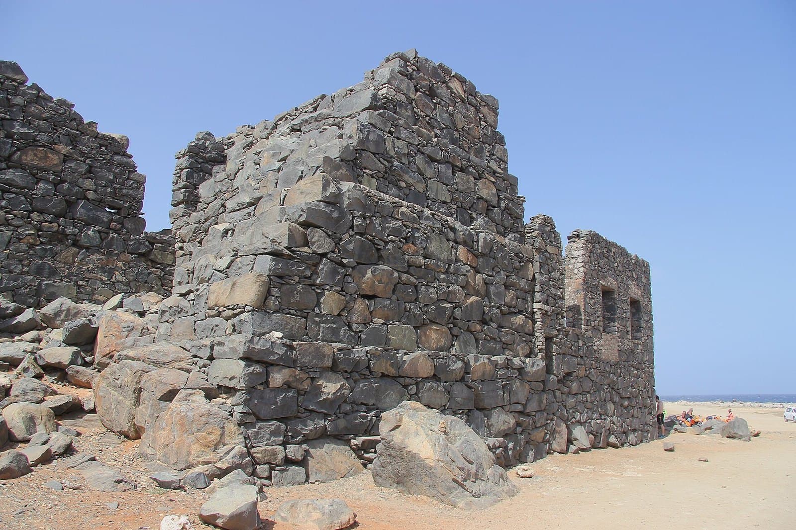 The dark fieldstone walls of the ruined Bushiribana gold smelter on Aruba's north coast, standing on open sand and rock with the sea visible beyond
