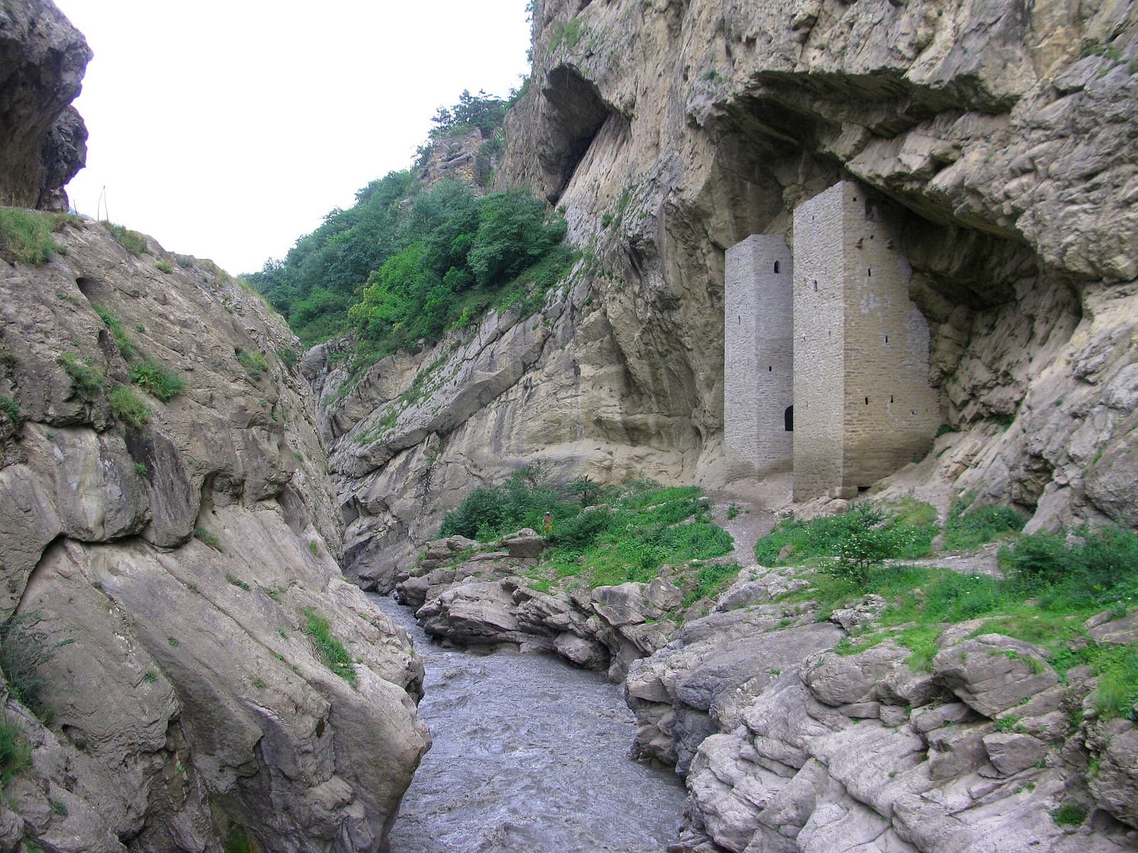 Two medieval stone battle towers standing under an overhanging limestone cliff above the Argun river at Ushkaloy, the pale rock face rising steeply on both sides of the gorge