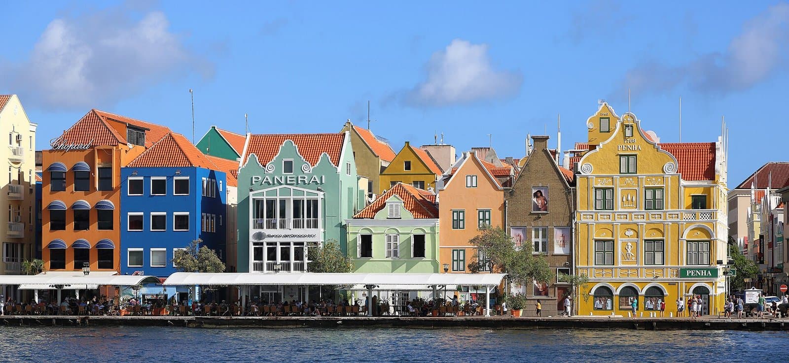 The Handelskade waterfront in Punda, Willemstad, a row of Dutch colonial merchant houses painted orange, blue, mint green, ochre and yellow with stepped gables and red tiled roofs, including the yellow Penha Building dated 1708