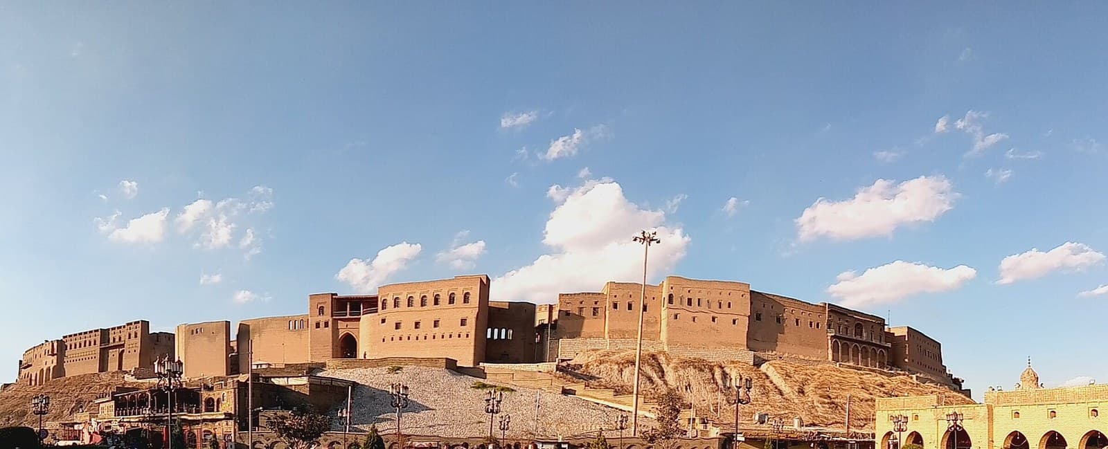 The Erbil Citadel seen from the plaza below, a long band of pale brick houses and an arched gatehouse along the top of a steep earthen mound, under a blue sky with scattered cloud