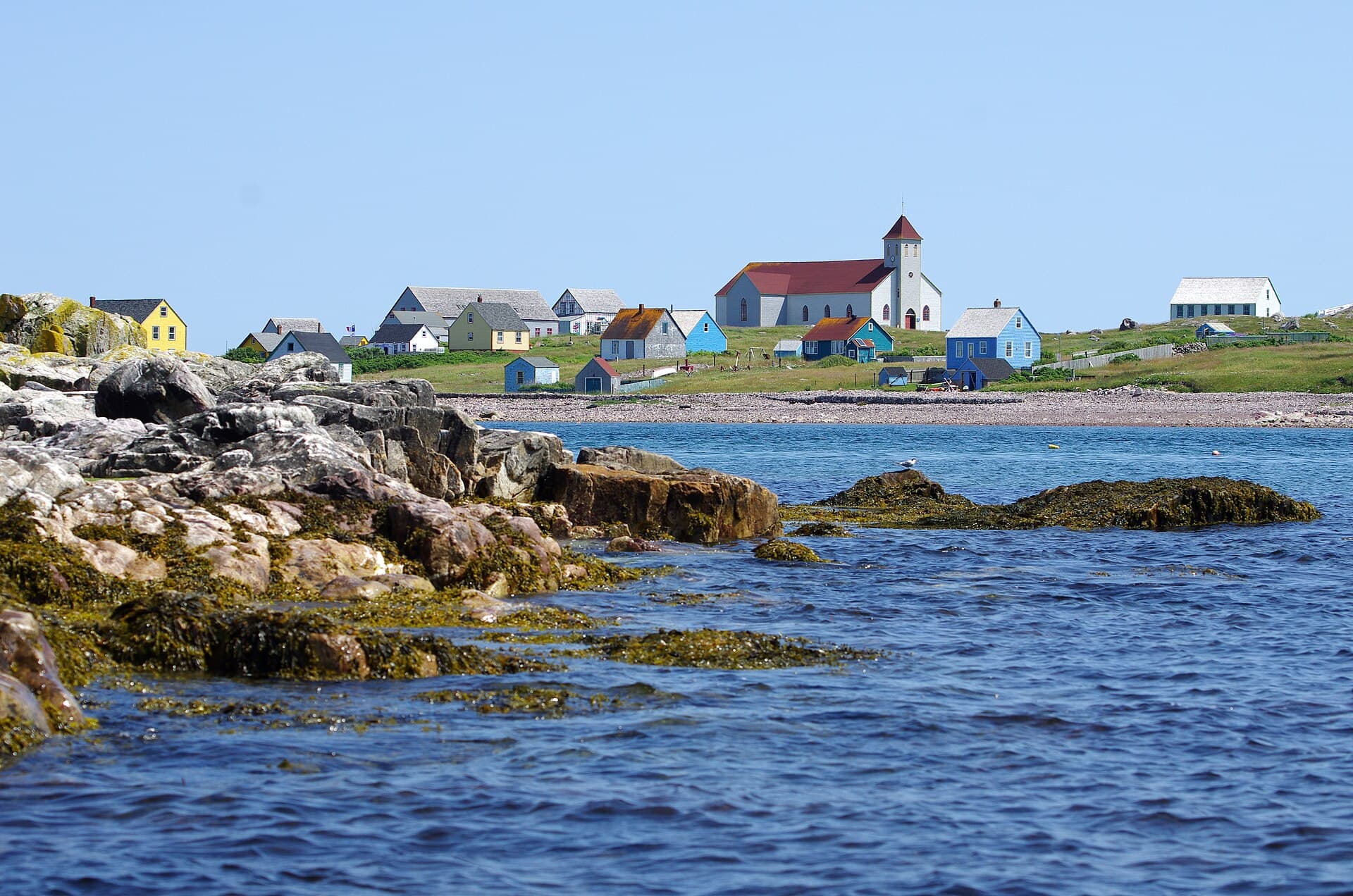 The abandoned village on Île aux Marins in Saint Pierre and Miquelon seen across the water, its brightly painted wooden houses and the white church of Notre-Dame-des-Marins standing on low green ground above a shingle beach