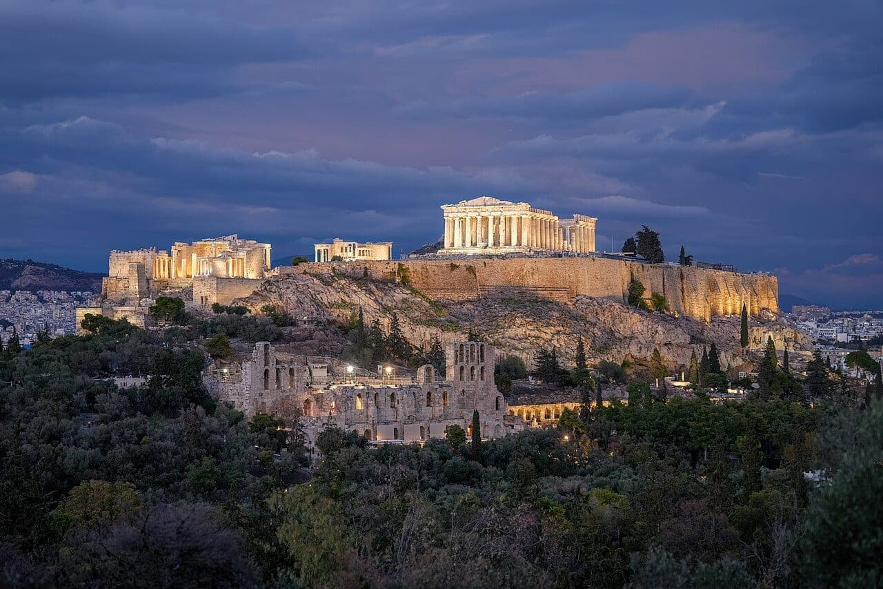 The floodlit Parthenon and marble temples of the Acropolis of Athens glowing above the city against a dark night sky.