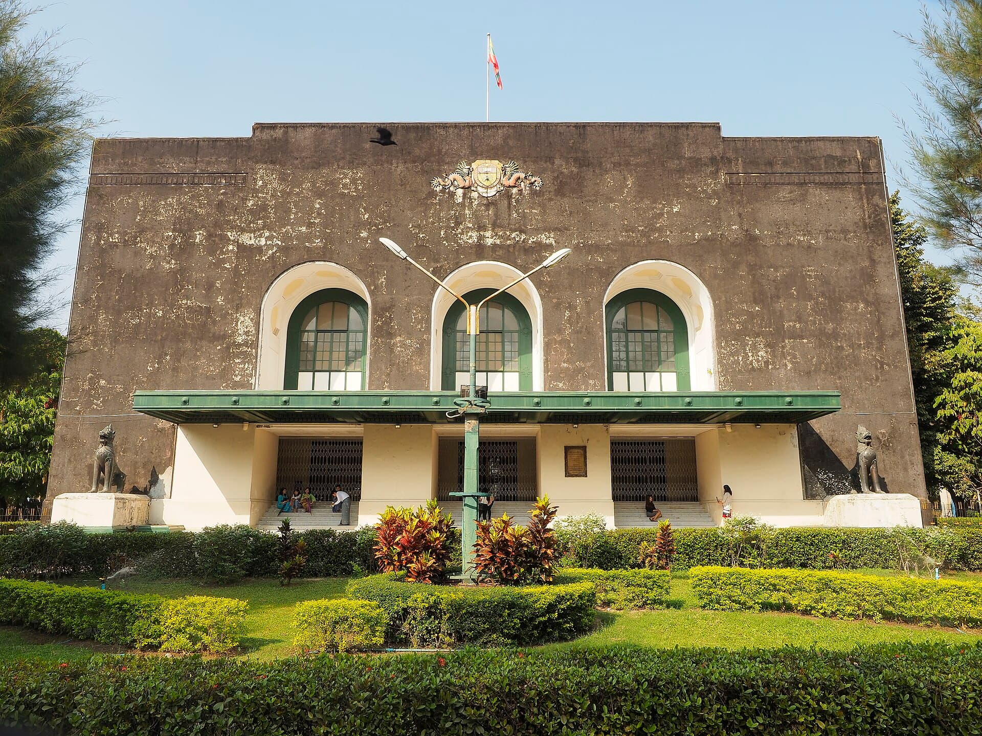 The weathered front of the Convocation Hall at the University of Yangon, with three tall arched windows and chinthe lion figures on plinths at either end